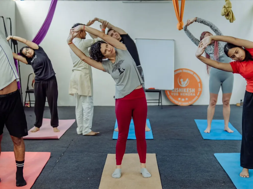 Indian instructor leading a standing side bend in the yoga hall at Rishikesh Yog Kendra