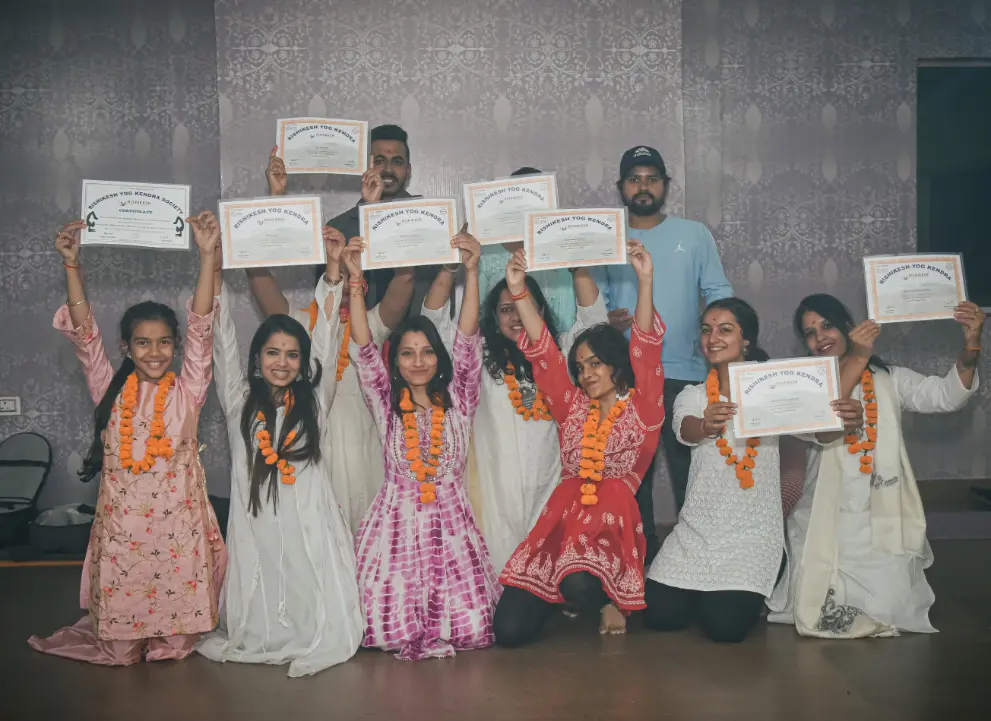 Students holding their Rishikesh Yog Kendra certificates overhead in the practice hall, marigold garlands around their necks