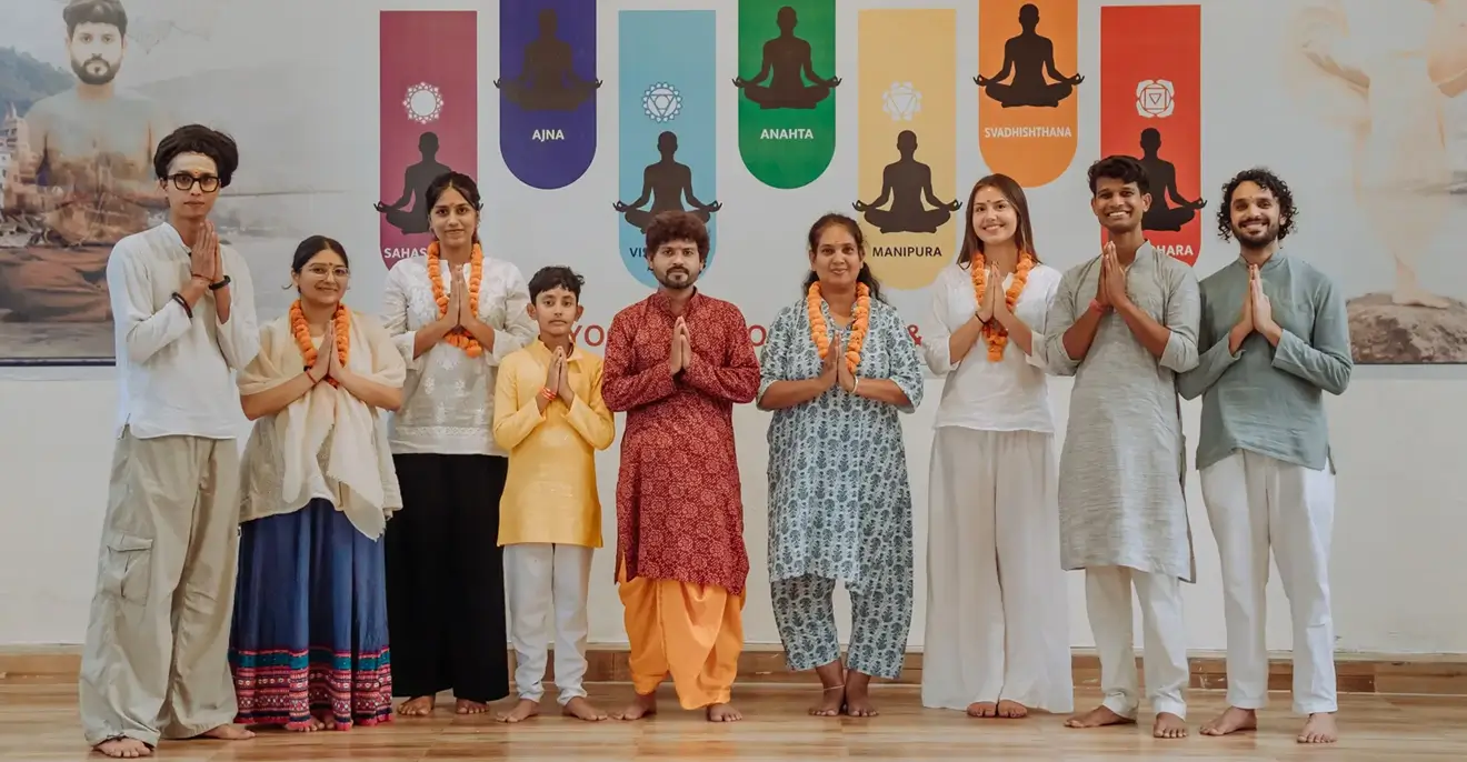 Teachers and students at Rishikesh Yog Kendra standing together in namaste in front of the chakra banner in the yoga shala