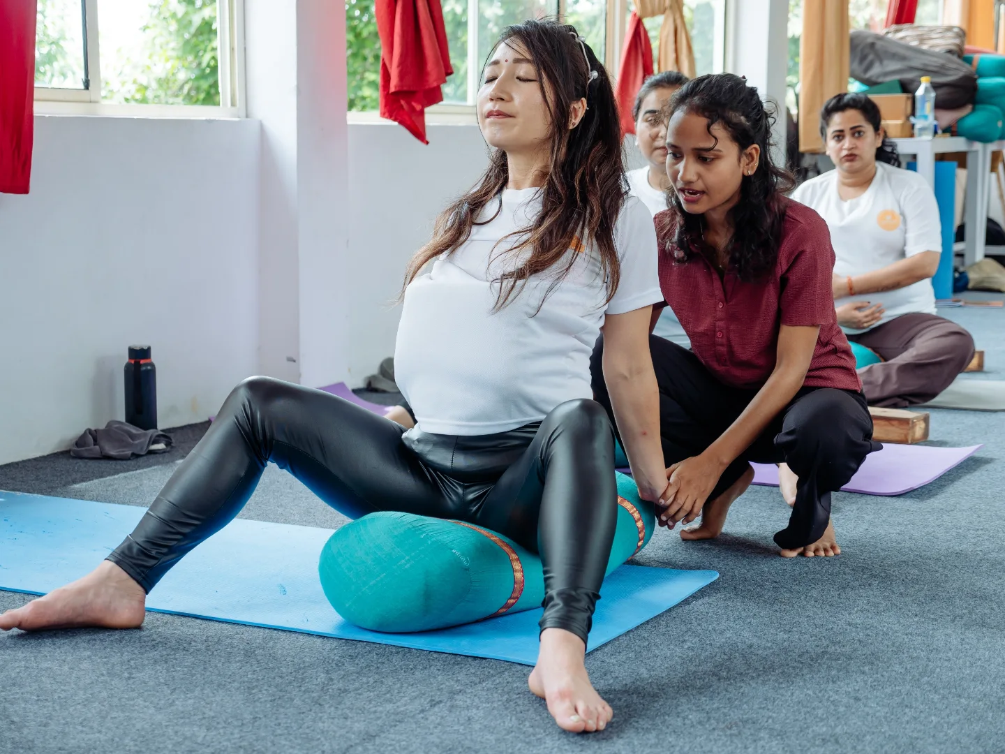 Teacher giving a hands-on adjustment to a pregnant student resting on a bolster during prenatal yoga class in Rishikesh