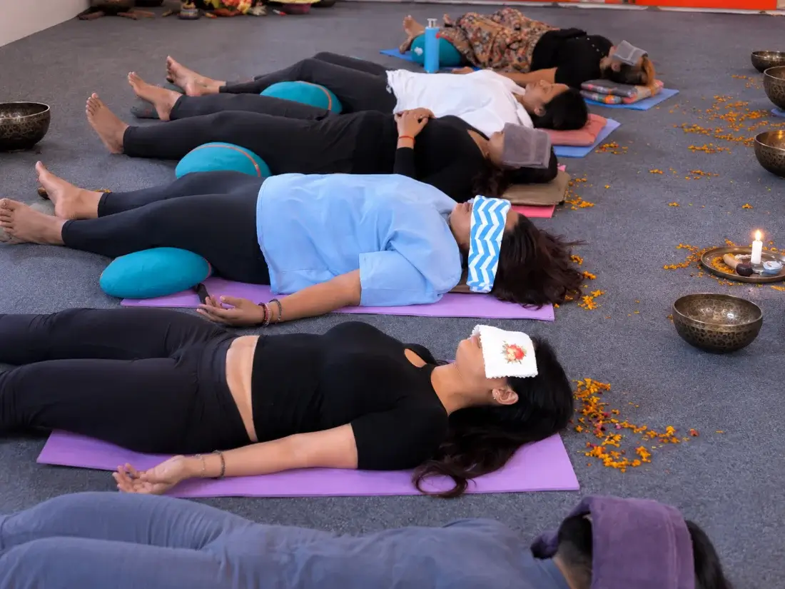 Students resting in savasana with bolsters and eye covers as bowls and a candle are set around the room