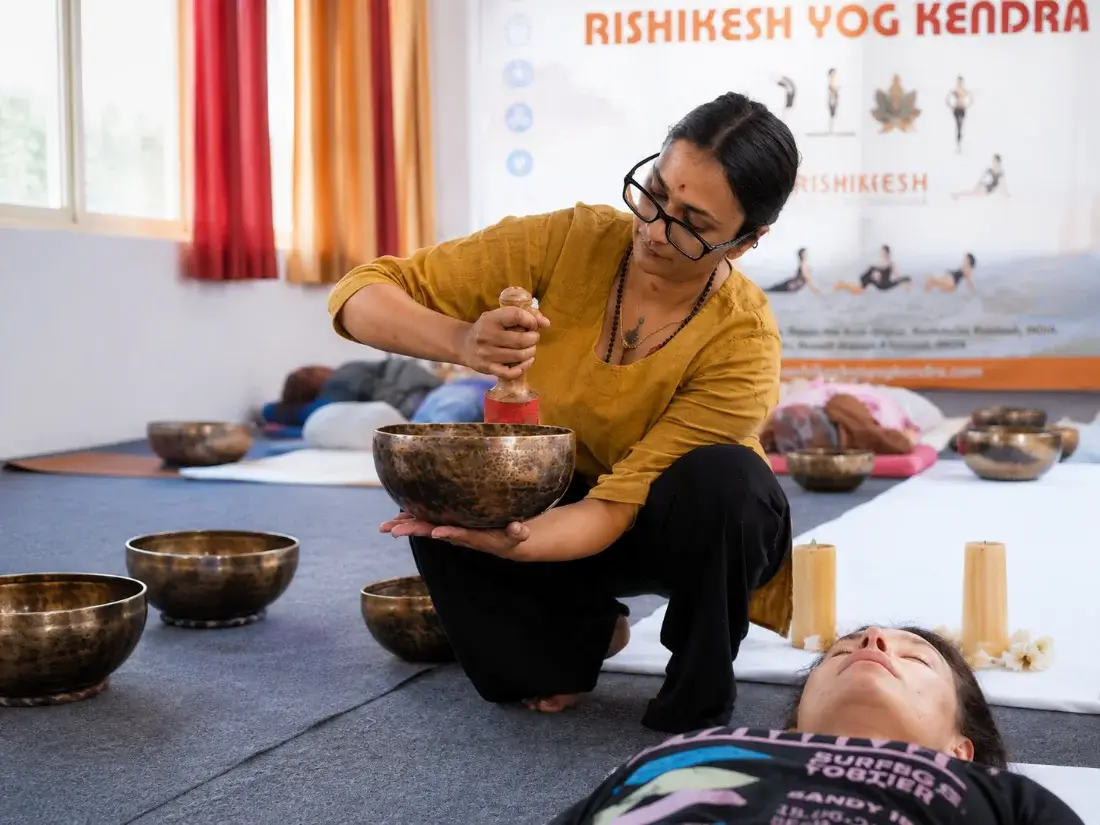 Teacher sounding a Tibetan singing bowl over a resting student during a sound healing session in Rishikesh