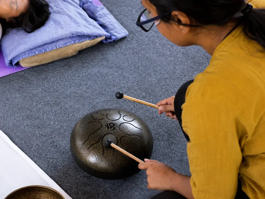 Hands striking a steel tongue drum beside a resting student